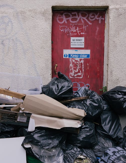 A waste collection worker operating a large red rubbish truck parked on the side of a street during daytime. The worker, dressed in a high-visibility yellow safety vest over a red jacket, is standing 