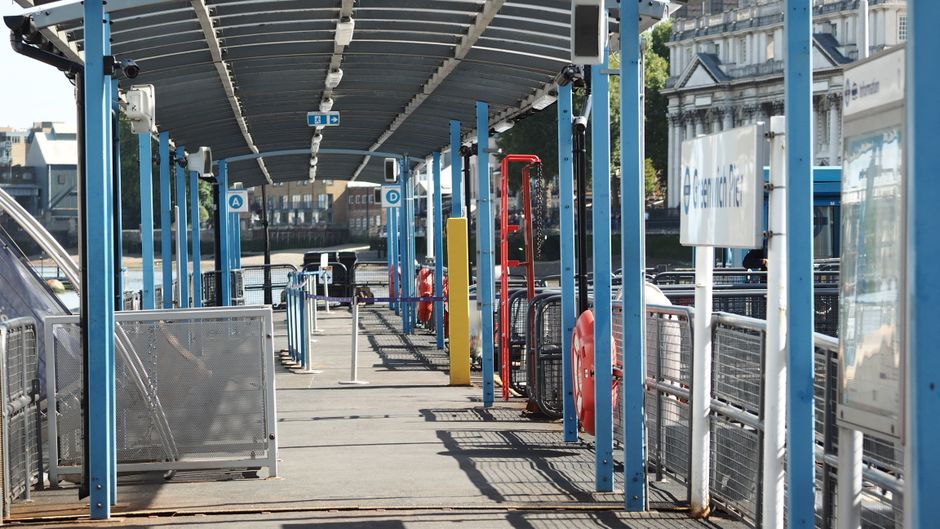 A covered outdoor area at a Woolwich Ferry rubbish drop-off point featuring blue metal posts supporting a corrugated roof. The space includes various waste management equipment such as red and orange